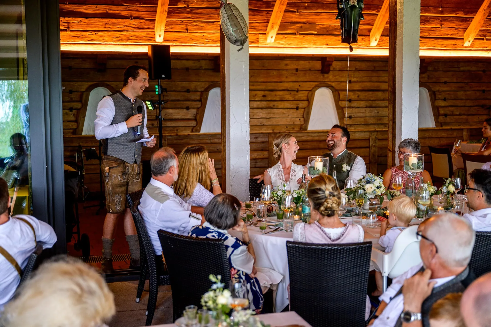 Hochzeitsfeier mit Rede, Brautpaar lacht am Tisch, Hochzeitsreportage in traditioneller Umgebung.