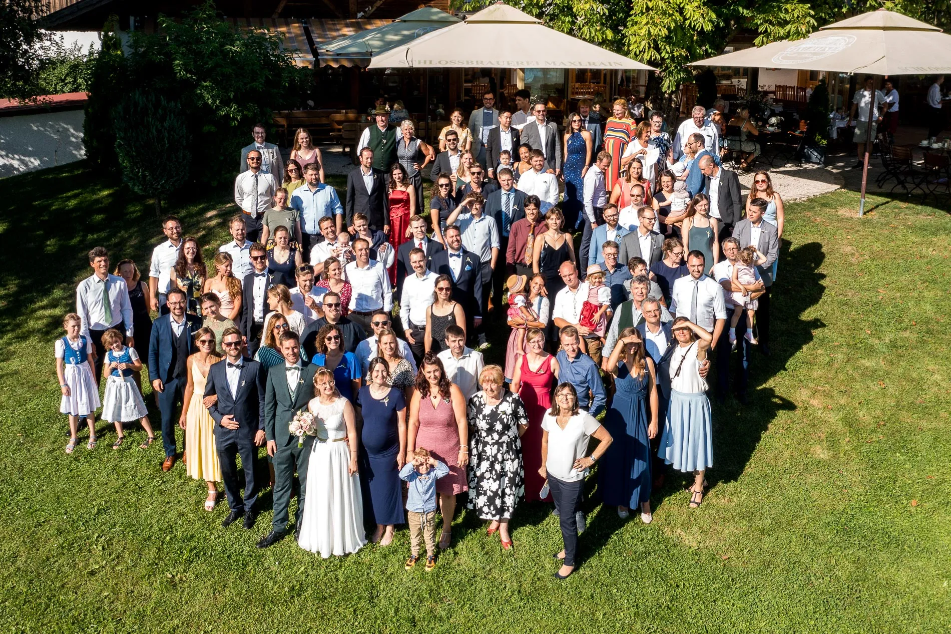 Hochzeitsfeier im Landgut Kugler-Alm, Ebersberg, mit Hochzeitsgesellschaft im Freien unter Sonnenschein. Gruppenfoto mit Drohne
